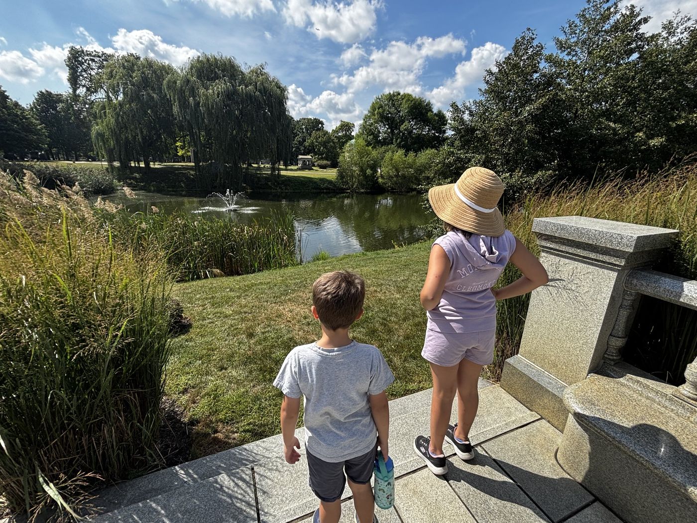 Two kids standing on a stone path, looking out over a pond with a fountain